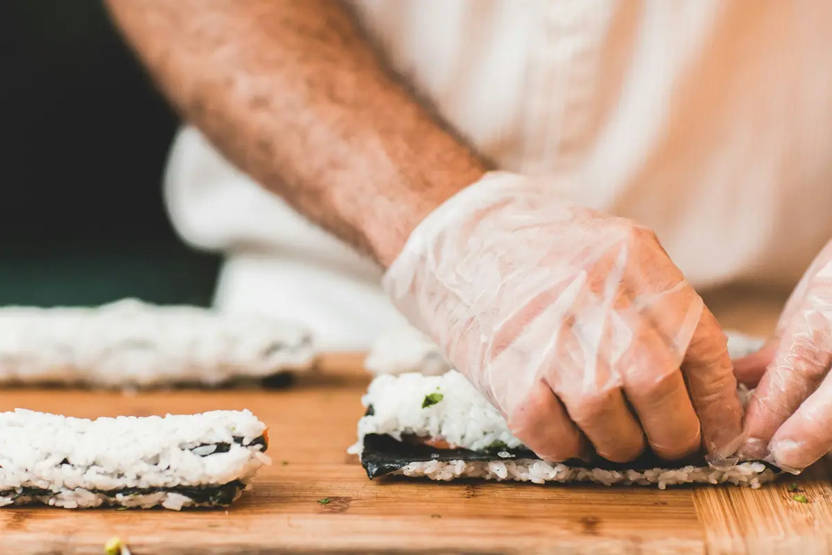 Gloved hands rolling rice and seaweed to make sushi